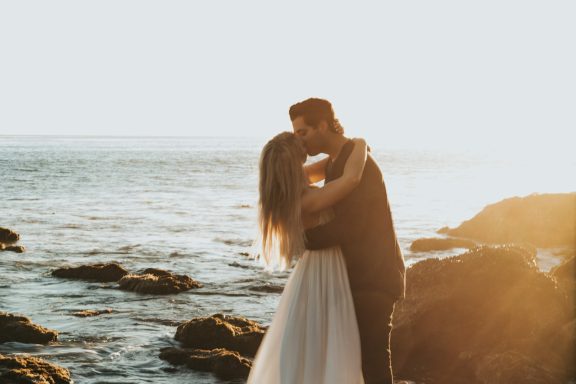 A couple in wedding attire embraces on a rocky beach at sunset.