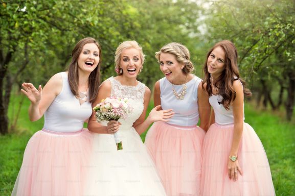 Four women in pink skirts and white tops, celebrating joyfully outdoors, with a bride in the centre.