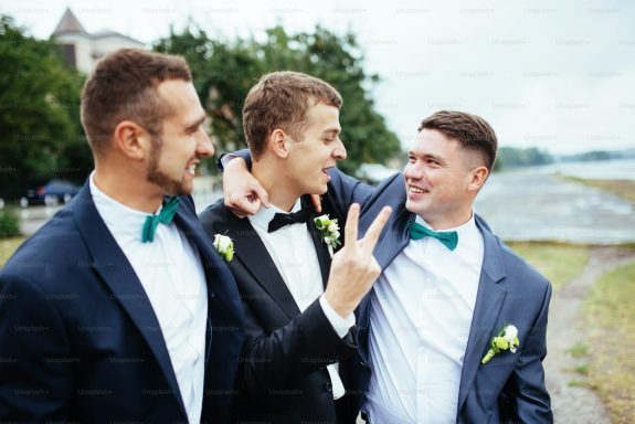 Three men in formal wear, smiling and posing together by a river.