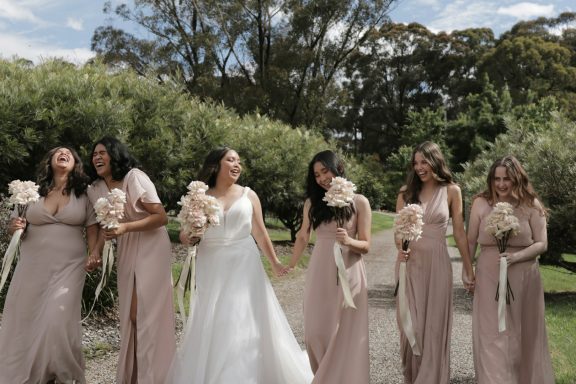 A bride and five bridesmaids in elegant dresses holding flowers, smiling outdoors.