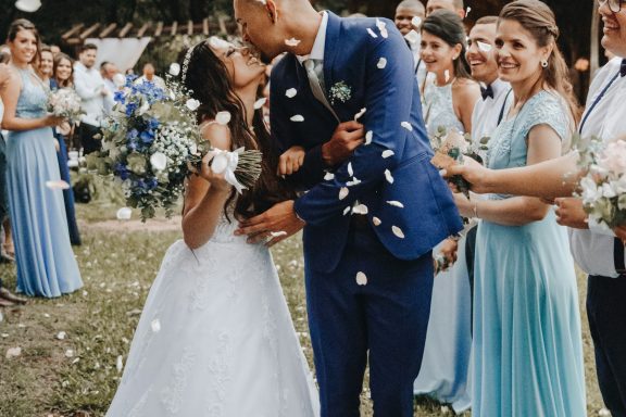 Bride and groom kiss surrounded by smiling wedding guests tossing confetti.