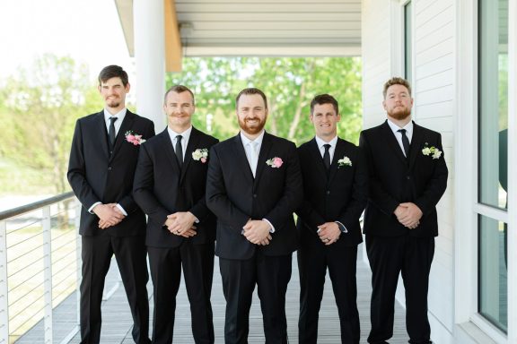 Five men in black suits stand together, smiling, outdoors near a building.