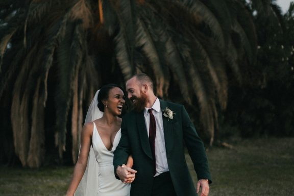A laughing couple in wedding attire walking hand in hand near palm trees.
