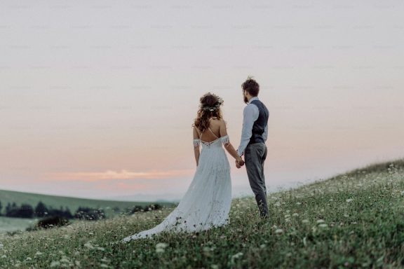 A couple holding hands, standing in a field at sunset, dressed in wedding attire.