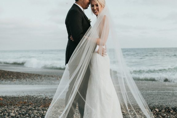 Bride and groom in elegant attire standing by the beach, veil gently blowing in the wind.
