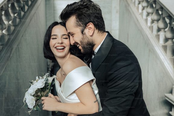 A joyful couple in formal wedding attire, laughing together on a staircase.