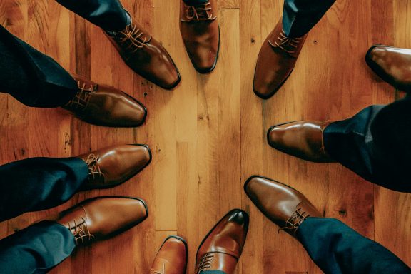 A circle of feet wearing various styles of shoes on a wooden floor.