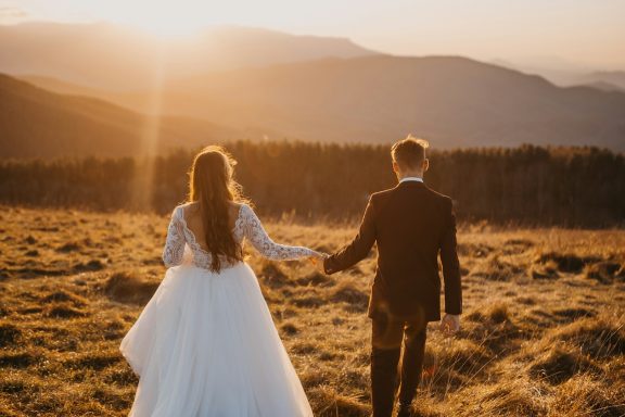 A couple holds hands, walking together towards a sunset over the mountains.